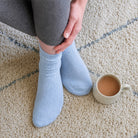 A person wearing light blue ribbed crew socks sitting on a beige carpet next to a ceramic mug of coffee.