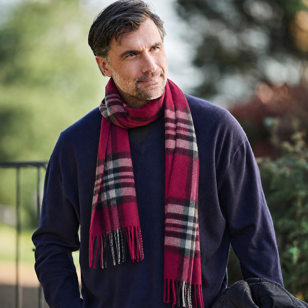 A man wearing a navy blue sweater and a red and black plaid scarf, photographed outdoors with a blurred green background.
