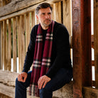 A man wearing a dark navy long-sleeve shirt and blue jeans sits on a rustic wooden bench, wrapped in a burgundy plaid scarf with white and black stripes.