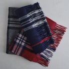 Three folded plaid and striped wool scarves with fringed edges, stacked on a light gray surface. The top scarf is navy blue with white and gray stripes, the middle scarf is red and pink plaid, and the bottom scarf is dark gray and black plaid.