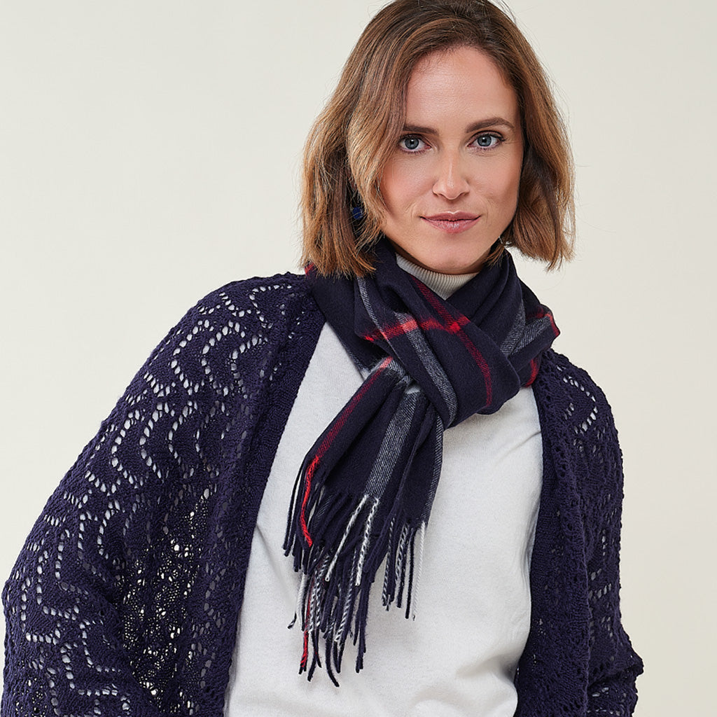 A woman wearing a navy blue open-knit cardigan and a dark navy plaid scarf with red and gray stripes, layered over a white turtleneck sweater, against a plain light beige background.
