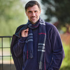A smiling man outdoors wearing a navy blue sweater and a thick navy scarf with gray, white, and red plaid stripes. He has a dark jacket draped over his shoulder, with a blurred green and brown background.