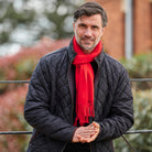 A man wearing a black quilted jacket and a bright red scarf, standing outdoors with his hands clasped in front of a blurred background.