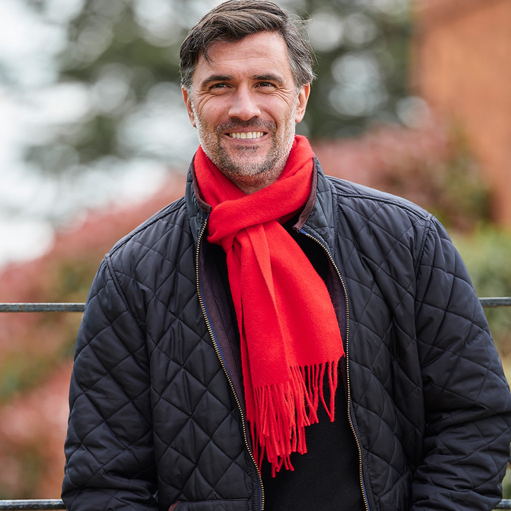 A smiling man wearing a bright red knit scarf and a black quilted jacket, photographed outdoors with a blurred background.