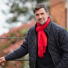 A man wearing a black quilted jacket and a bright red knit scarf, standing outdoors with a blurred background of trees and a brick wall.