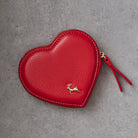 A red heart-shaped leather coin purse with a gold deer logo and a matching red leather zipper pull, shown on a textured gray background.