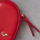 A close-up of a red textured leather coin purse with a gold deer logo and a matching red leather zipper pull on a gray background.