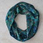 A peacock feather print infinity scarf in shades of teal, blue, and brown, displayed in a loop on a light gray textured surface.