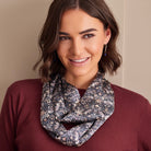 A smiling woman with shoulder-length brown hair models a patterned infinity scarf. The scarf features a detailed floral and paisley design in navy blue, beige, and white.
