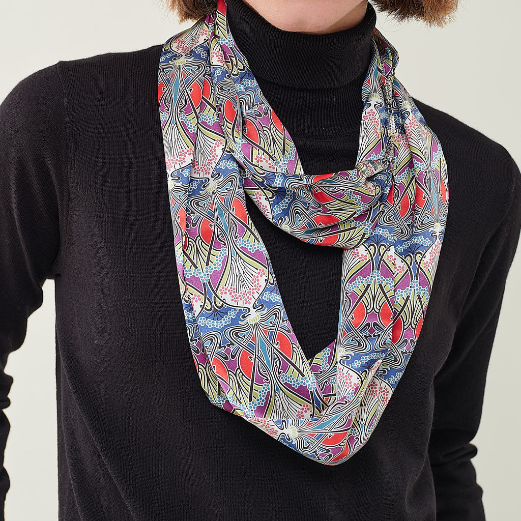 A multicolor abstract patterned scarf with red, blue, green, and purple swirls, worn as a loop necklace over a black turtleneck sweater.