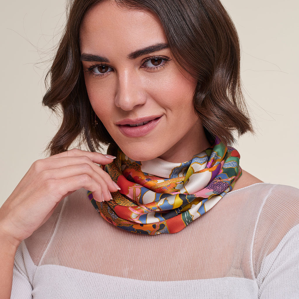 A woman with short brown hair models a colorful, patterned infinity scarf wrapped around her neck, wearing a sheer white top against a light beige background.