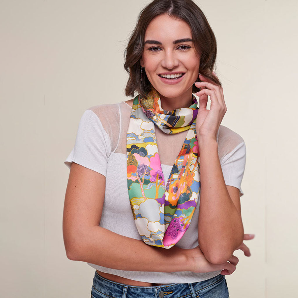 A woman wearing a white short-sleeve top and blue jeans, smiling and posing with a colorful abstract-patterned scarf draped around her neck, set against a plain beige background.
