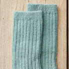 A pair of ribbed knit socks in a dusty sage green color, displayed on a light wood background.
