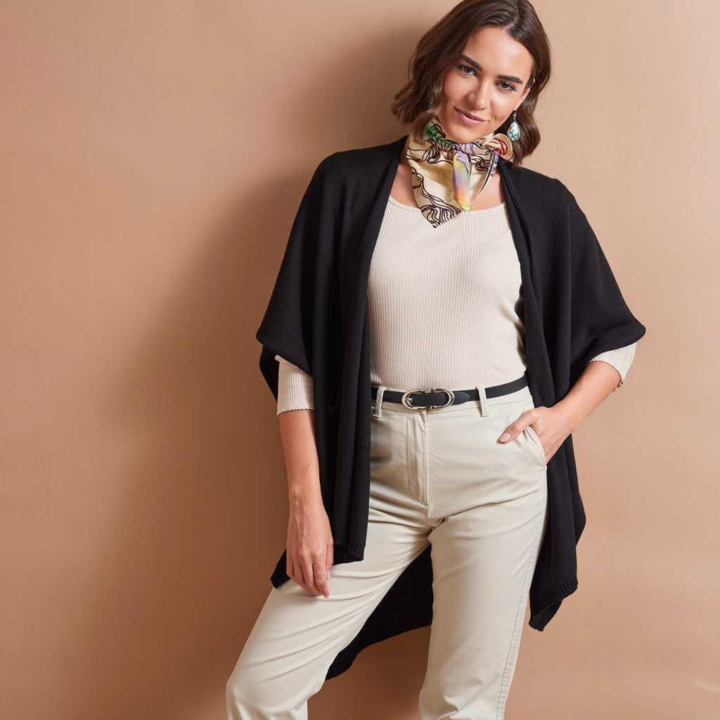 A woman models a black open-front drape cardigan over a cream ribbed top and cream trousers, accessorized with a patterned neck scarf, against a solid tan studio background.
