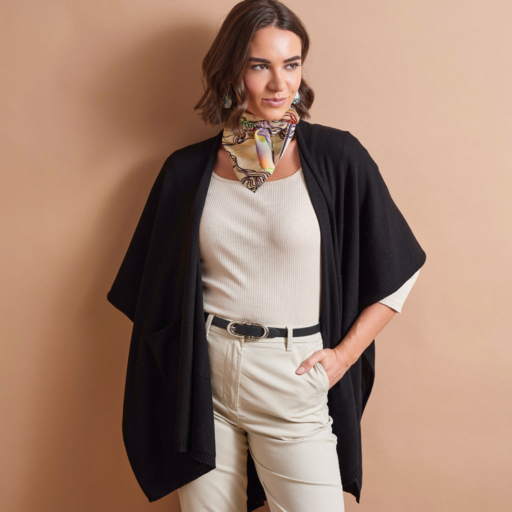 A woman models a black open-front cape cardigan over a cream ribbed top and cream pants, accessorized with a multicolor patterned neck scarf, against a solid beige studio background.