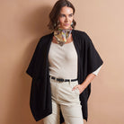 A woman models a black open-front cape cardigan over a cream ribbed top and cream pants, accessorized with a multicolor patterned neck scarf, against a solid beige studio background.