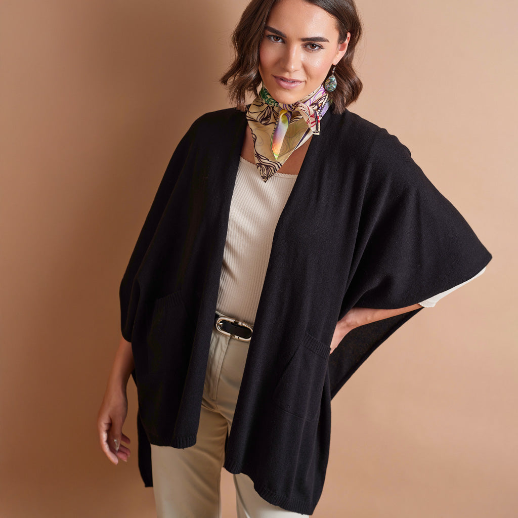 A woman models a black open-front poncho-style cardigan over a cream ribbed top and beige pants, accessorized with a multicolor patterned neck scarf, against a solid warm beige studio background.