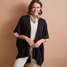 A woman models a black open-front lightweight cardigan or poncho over a cream ribbed top and cream pants, accessorized with a patterned neck scarf, against a solid warm beige studio background.