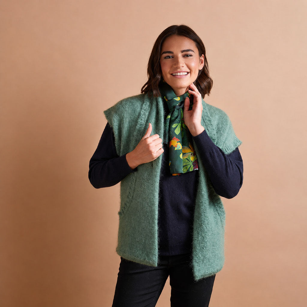 A woman modeling a fuzzy sage green knit vest over a dark navy long-sleeve top and black pants, accessorized with a green floral neck scarf, against a solid warm tan studio background.