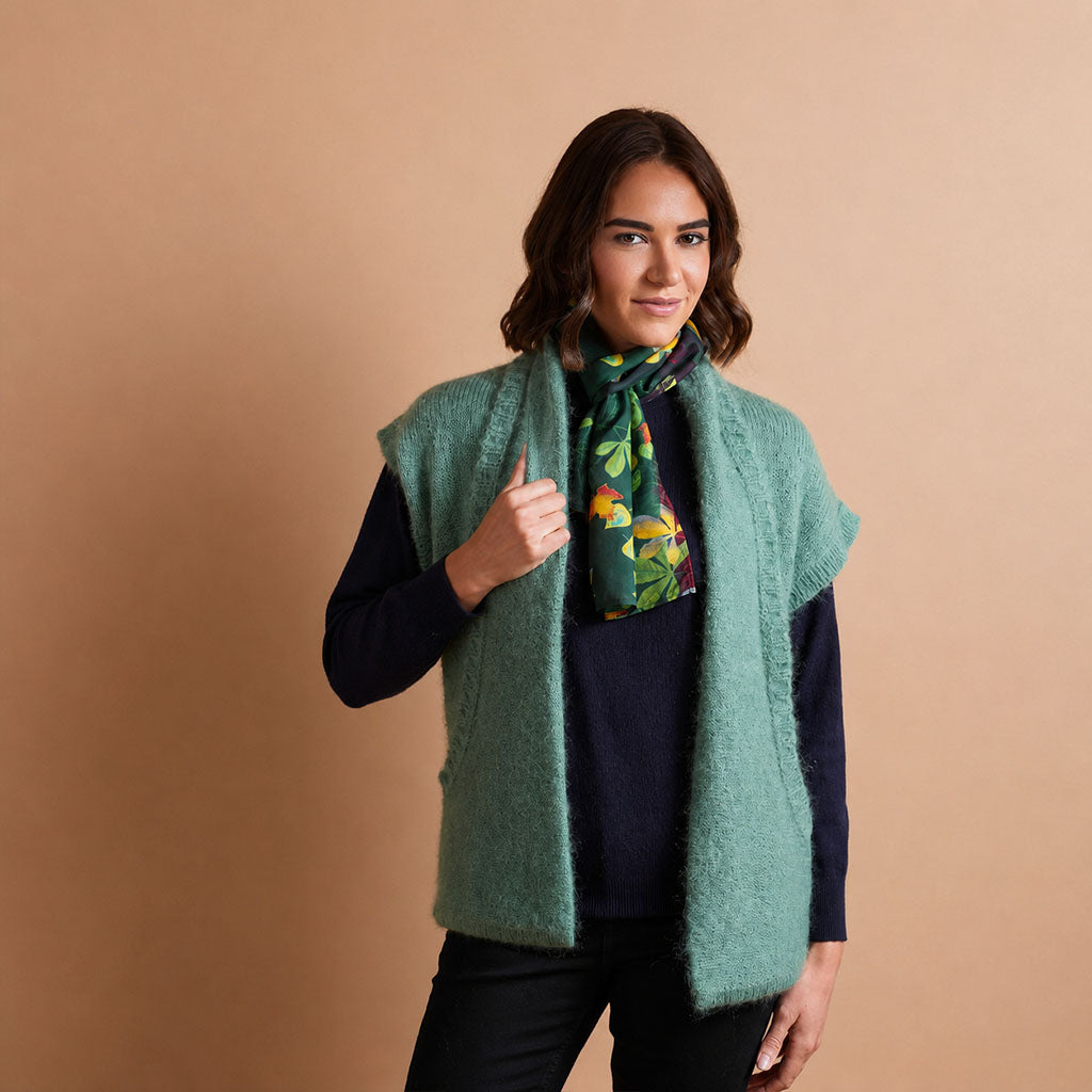 A woman modeling a fuzzy sage green sleeveless vest over a dark navy sweater, accessorized with a green and yellow patterned scarf, set against a solid warm beige background.