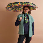 A woman holding a floral print umbrella, wearing a sage green fuzzy vest over a navy sweater, a green floral scarf, and black pants, set against a solid warm beige background.