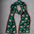 A green silk scarf with a repeating circular medallion pattern in red, white, and gold, finished with a red border, laid flat on a gray textured background.