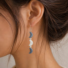 A close-up of a person's ear wearing a dangling earring with three stacked crescent moon charms in blue, white, and blue, suspended from a silver hook.