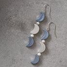 A pair of dangling crescent moon earrings with alternating light blue and white moon charms, separated by small white pearl beads, hanging from silver fishhook wires on a textured gray background.