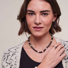 A woman with short brown hair models a beaded necklace with black, white, and pinkish-red beads, paired with a black top and a cream-colored crochet cardigan.