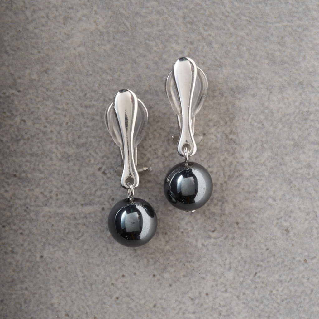 A pair of silver-toned drop earrings with polished oval posts and dark metallic spherical charms, displayed on a textured gray surface.