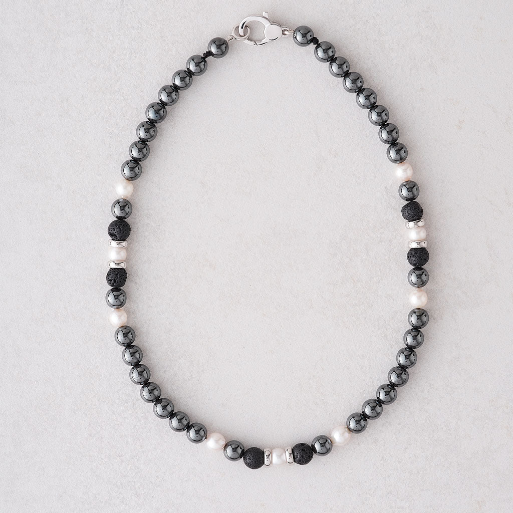 A beaded necklace featuring dark gray and black beads alternating with small white pearls, accented by silver spacer beads and a silver clasp, displayed on a light gray background.