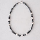 A beaded necklace featuring dark gray and black beads alternating with small white pearls, accented by silver spacer beads and a silver clasp, displayed on a light gray background.