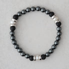 A beaded stretch bracelet featuring black and dark gray volcanic lava beads, white pearl-like beads, and silver-toned spacer beads, displayed on a light gray textured background.