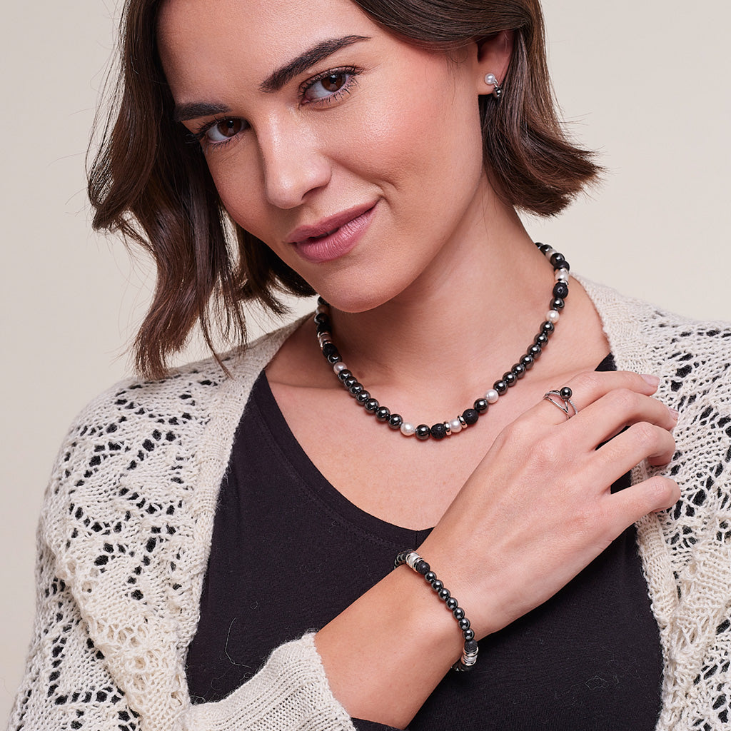 A woman wearing a black beaded necklace and matching beaded bracelet with silver-toned metal accents, styled with a black top and cream open-knit cardigan against a light beige background.