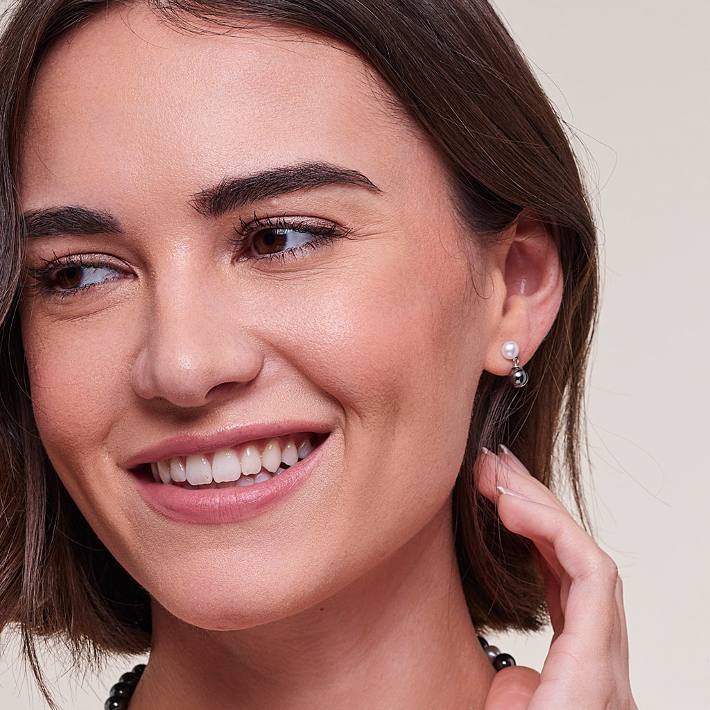 Close-up of a smiling woman wearing a small silver hoop earring with a single white pearl, against a soft beige background.