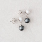A pair of silver stud earrings featuring a white pearl and a dark gray teardrop-shaped pearl, displayed on a light gray textured background.