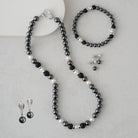 A jewelry set laid out on a light gray surface, including a beaded necklace, bracelet, earrings, and ring. The pieces feature glossy black beads, white pearl-like beads, and small silver-toned spacer beads, with silver-toned metal findings.