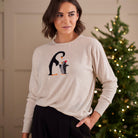 A woman wearing a light beige crewneck sweater with a penguin graphic, posed in front of a softly lit Christmas tree.