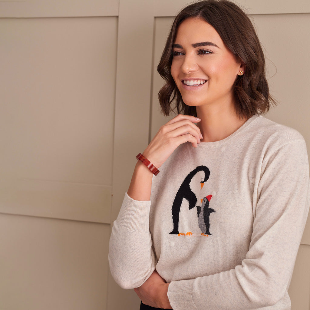 A woman wearing a light beige crewneck sweater with an embroidered penguin and chick design on the chest, posing against a neutral beige paneled wall.