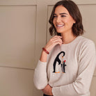 A woman wearing a light beige crewneck sweater with an embroidered penguin and chick design on the chest, posing against a neutral beige paneled wall.