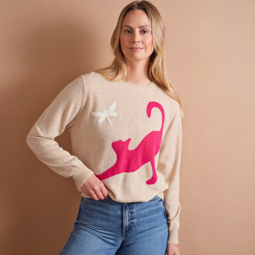 A woman wearing a beige crewneck sweater with a large hot pink cat silhouette and a small white butterfly graphic, paired with blue jeans, set against a solid warm beige background.