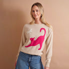 A woman wearing a beige crewneck sweater with a large hot pink cat graphic and a small white star detail, paired with blue jeans, set against a solid warm beige background.