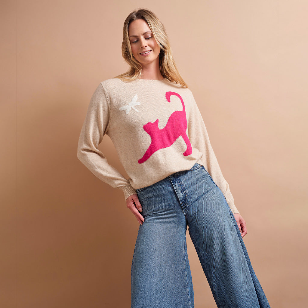 A woman modeling a beige knit sweater with a large pink cat graphic and a small white dragonfly motif, paired with wide-leg blue jeans against a solid warm beige studio background.
