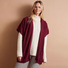 A woman modeling a burgundy open-front cardigan with cream cable-knit sleeves, worn over a cream turtleneck sweater and plaid trousers, against a solid beige background.