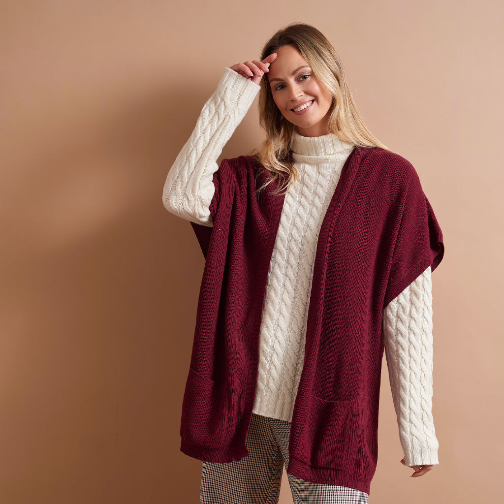 A woman modeling a cashmere burgundy open-front cardigan with cream cable-knit sleeves and a cream turtleneck, paired with gray plaid pants, against a solid warm beige background.