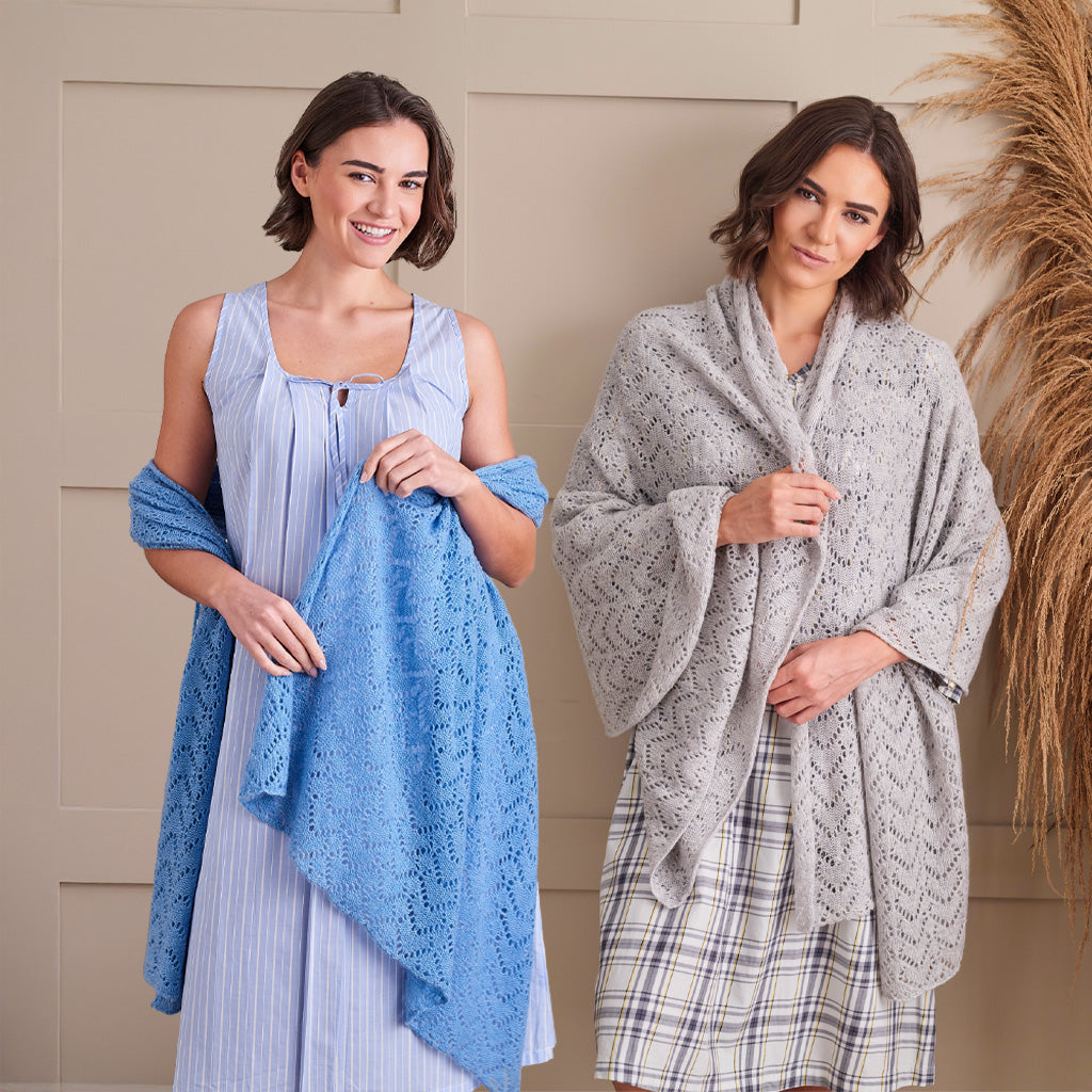Two women modeling knitted shawls in a studio setting. The woman on the left wears a light blue sleeveless dress and holds a light blue openwork shawl.