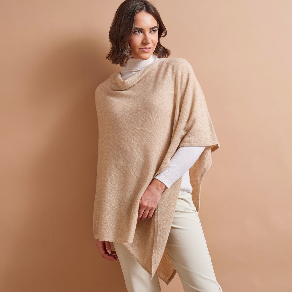 A woman modeling a light beige open-front knit poncho over a white mock-neck top and cream pants, set against a solid warm beige studio background.