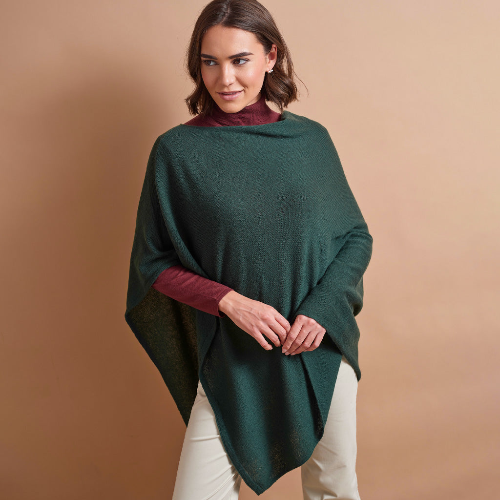 A woman modeling a dark forest green knit poncho layered over a burgundy mock neck top and cream pants, set against a solid warm tan studio background.