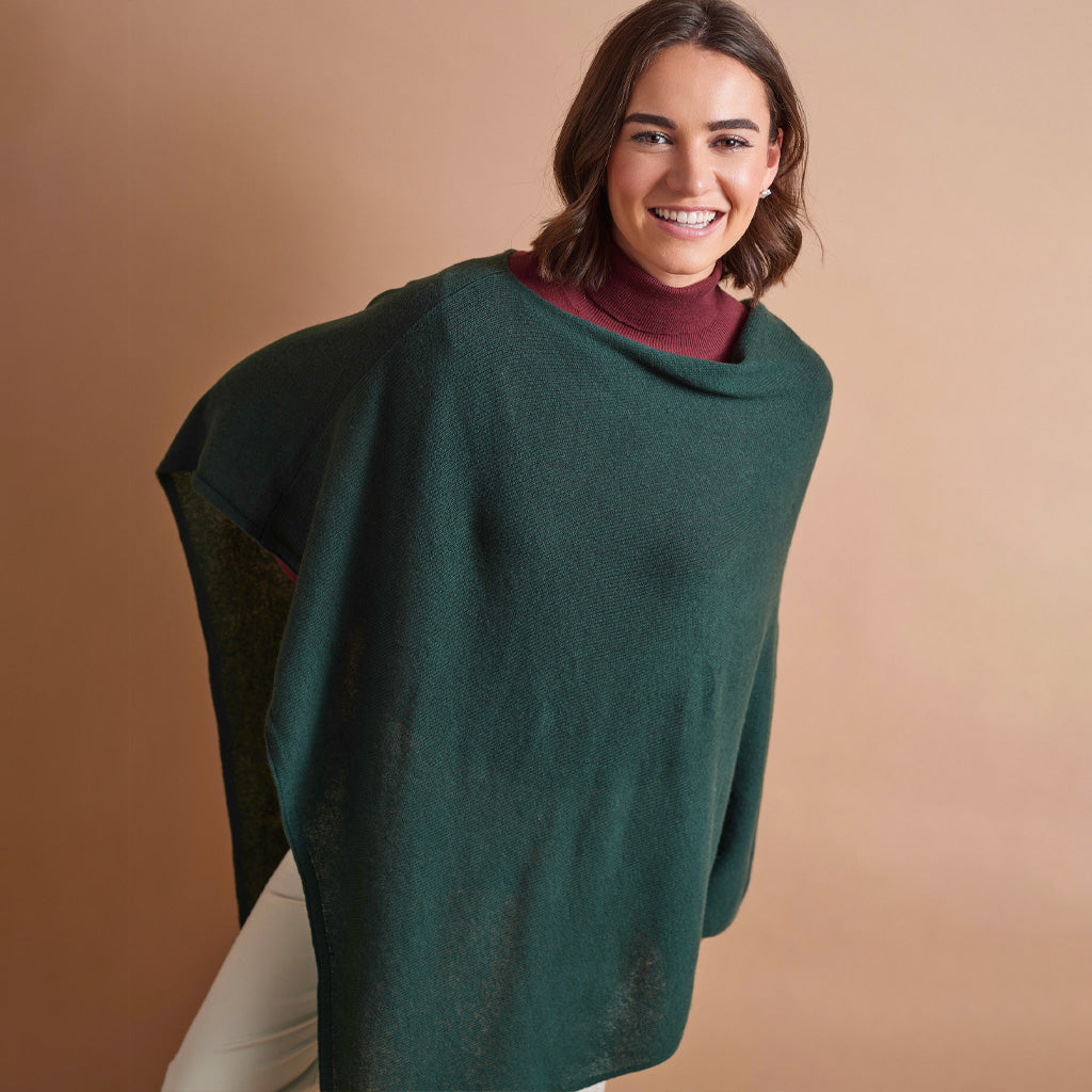 A woman wearing a dark green draped poncho over a burgundy turtleneck sweater and white pants, standing against a solid warm brown studio background.