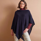 A woman modeling a dark navy blue knit poncho over a burgundy turtleneck and cream pants, set against a solid warm beige studio background.
